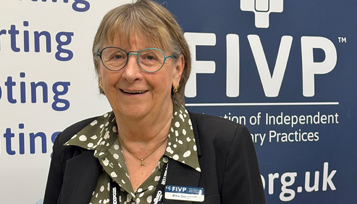 An image of Rita Dingwall, chair of FIVP. She wears glasses and a green blouse, and is standing in front of a blue FIVP logo.